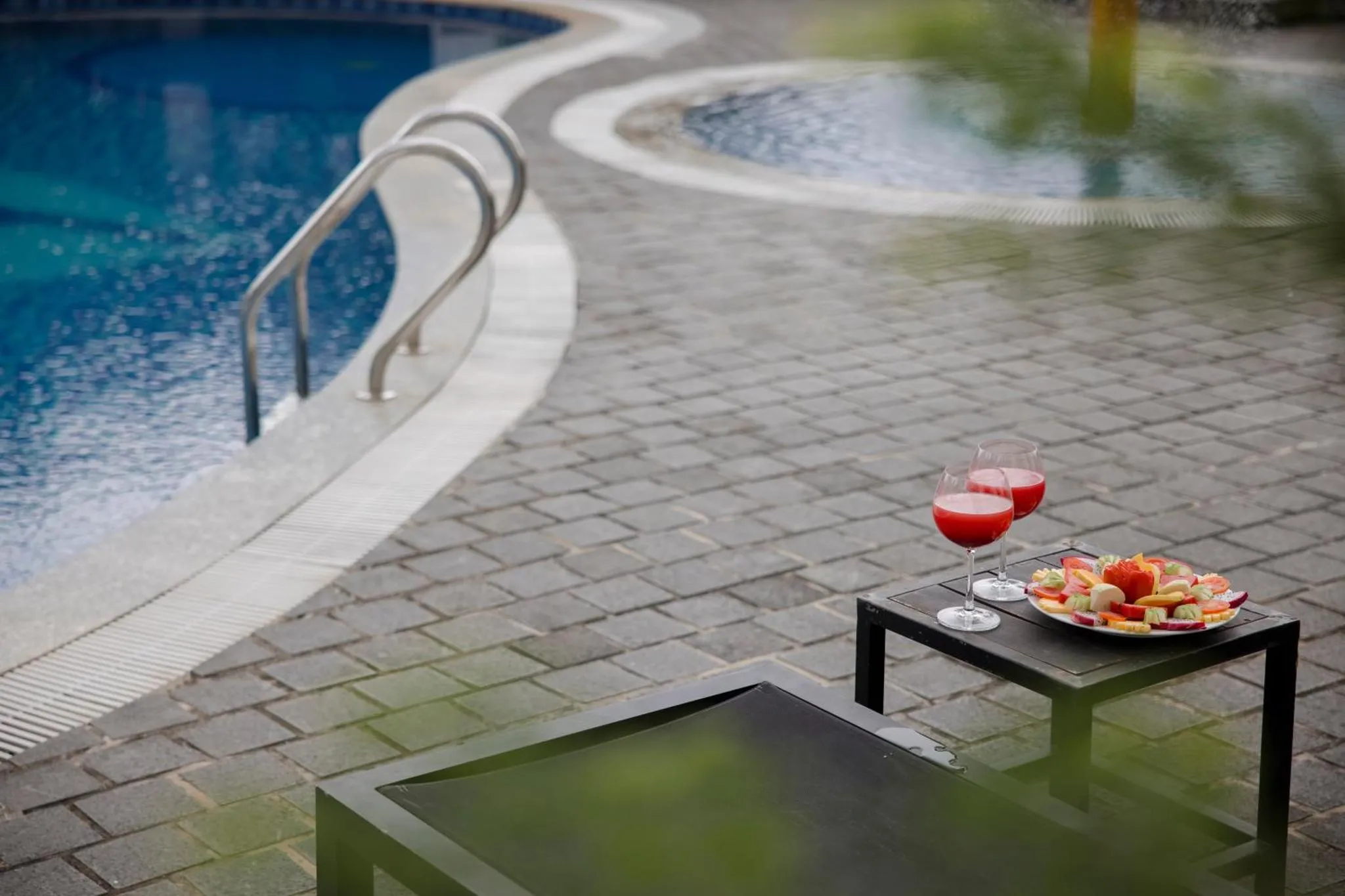 Deux verres et une assiette de fruits sont posés sur une table basse au bord d'une piscine.
