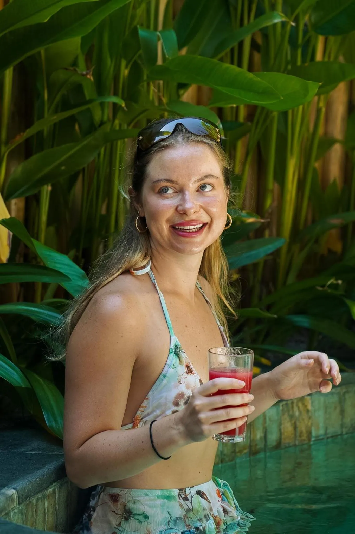 Une jeune femme souriante tient un verre coloré près d'une piscine.