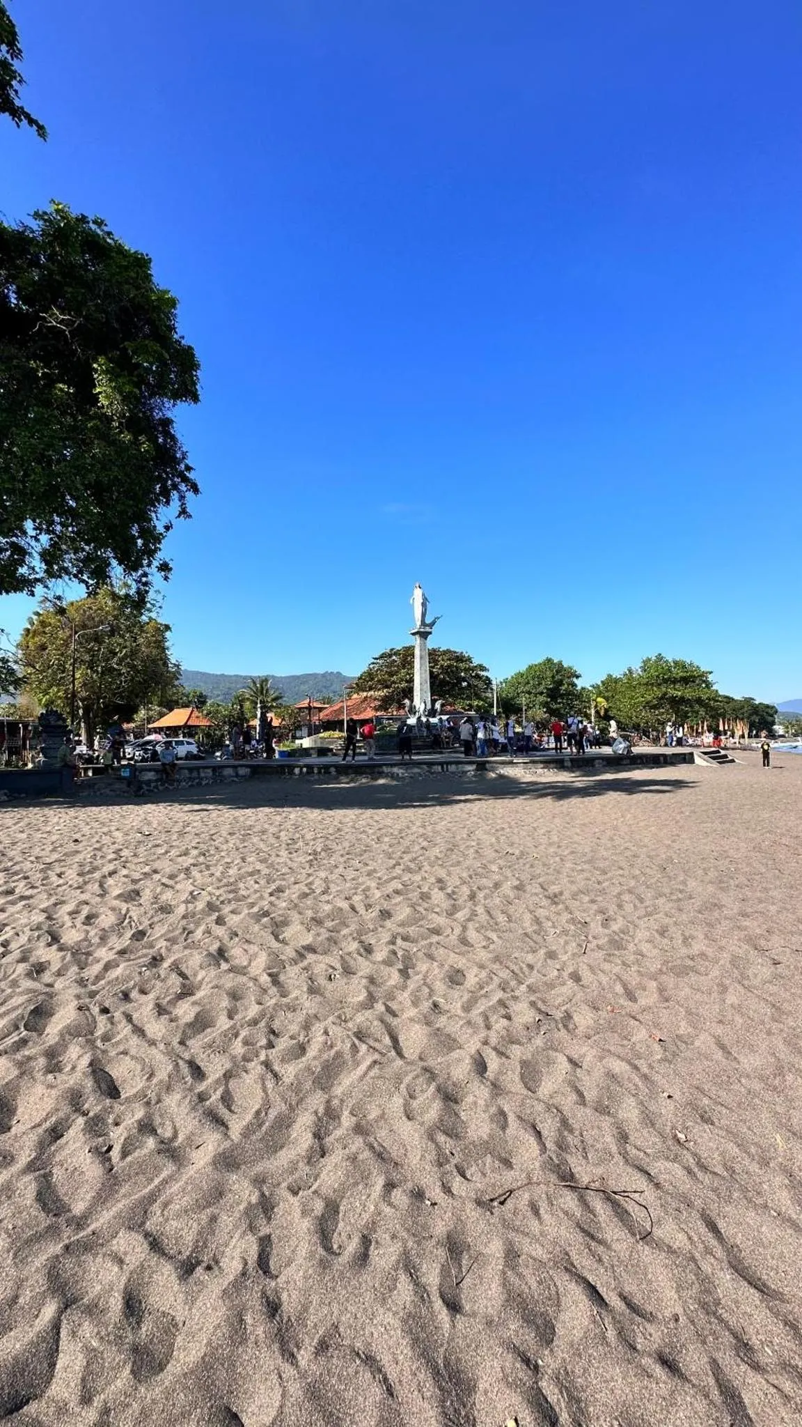 Plage de sable gris avec quelques personnes et des bâtiments en arrière-plan sous un ciel bleu.