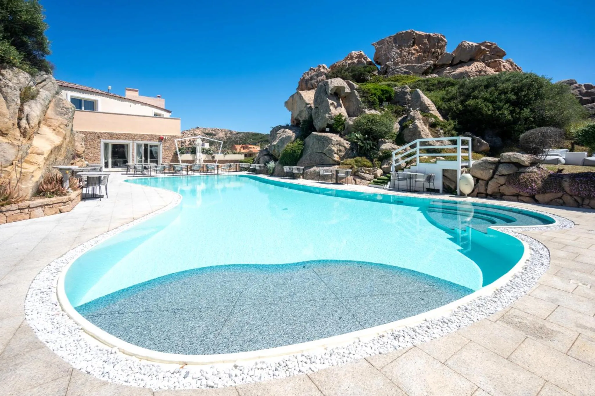 Une piscine bleue bordée d'une terrasse ensoleillée avec vue sur des rochers et la mer.