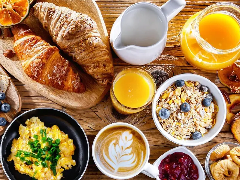 Un petit-déjeuner copieux avec viennoiseries, œufs brouillés, café et jus d'orange est présenté sur une table en bois.