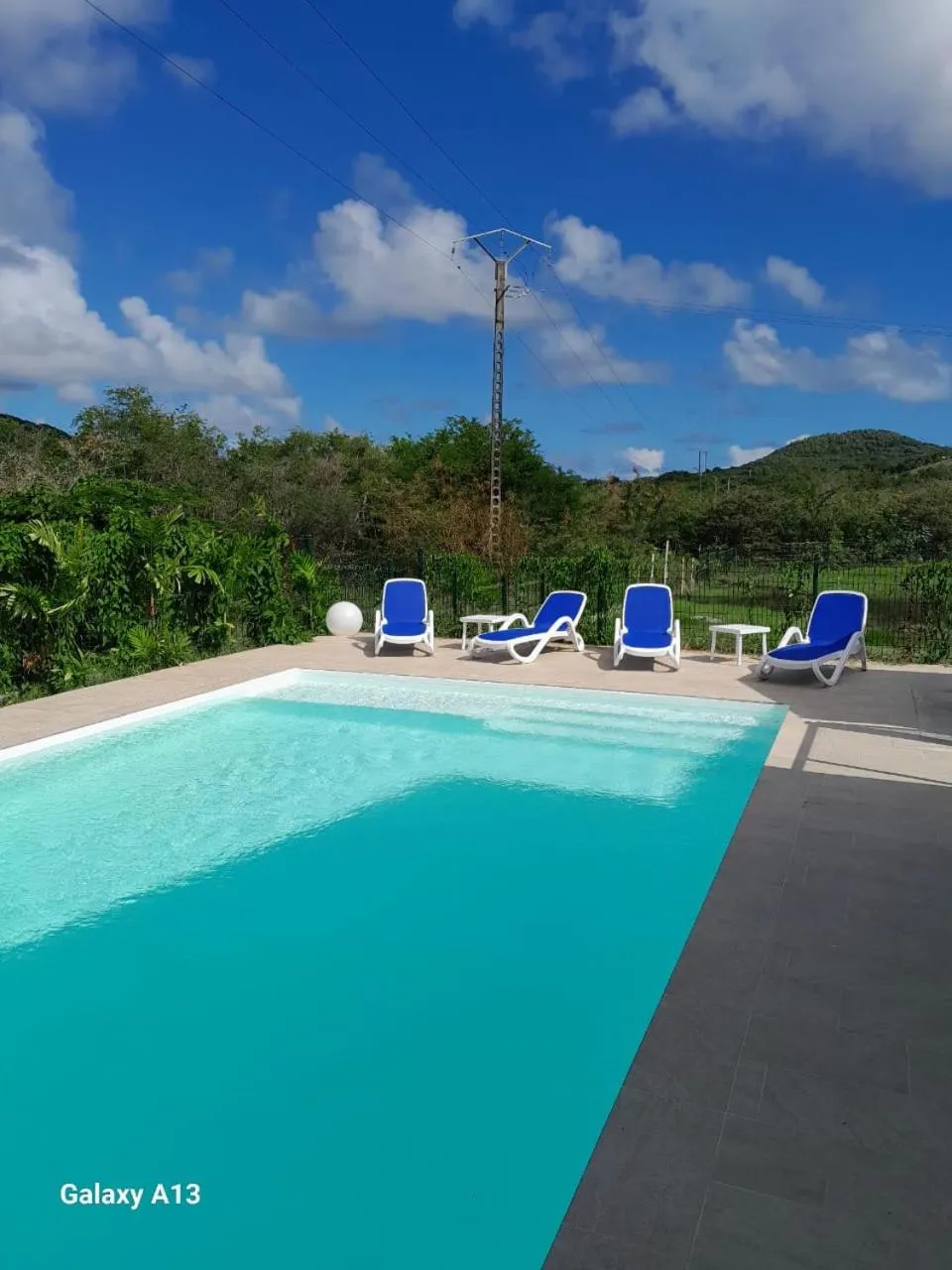 Piscine rectangulaire turquoise bordée d'une terrasse ensoleillée avec des transats bleus.
