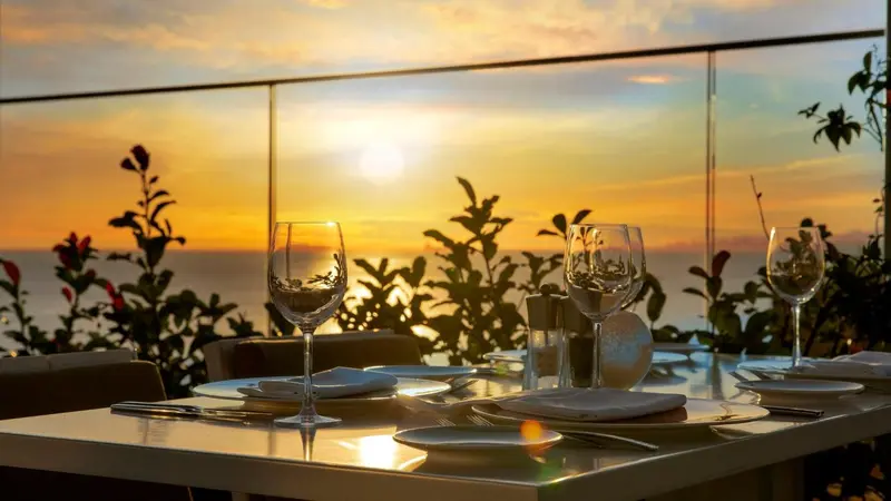 Une table dressée avec des verres et des couverts face à un coucher de soleil sur la mer.