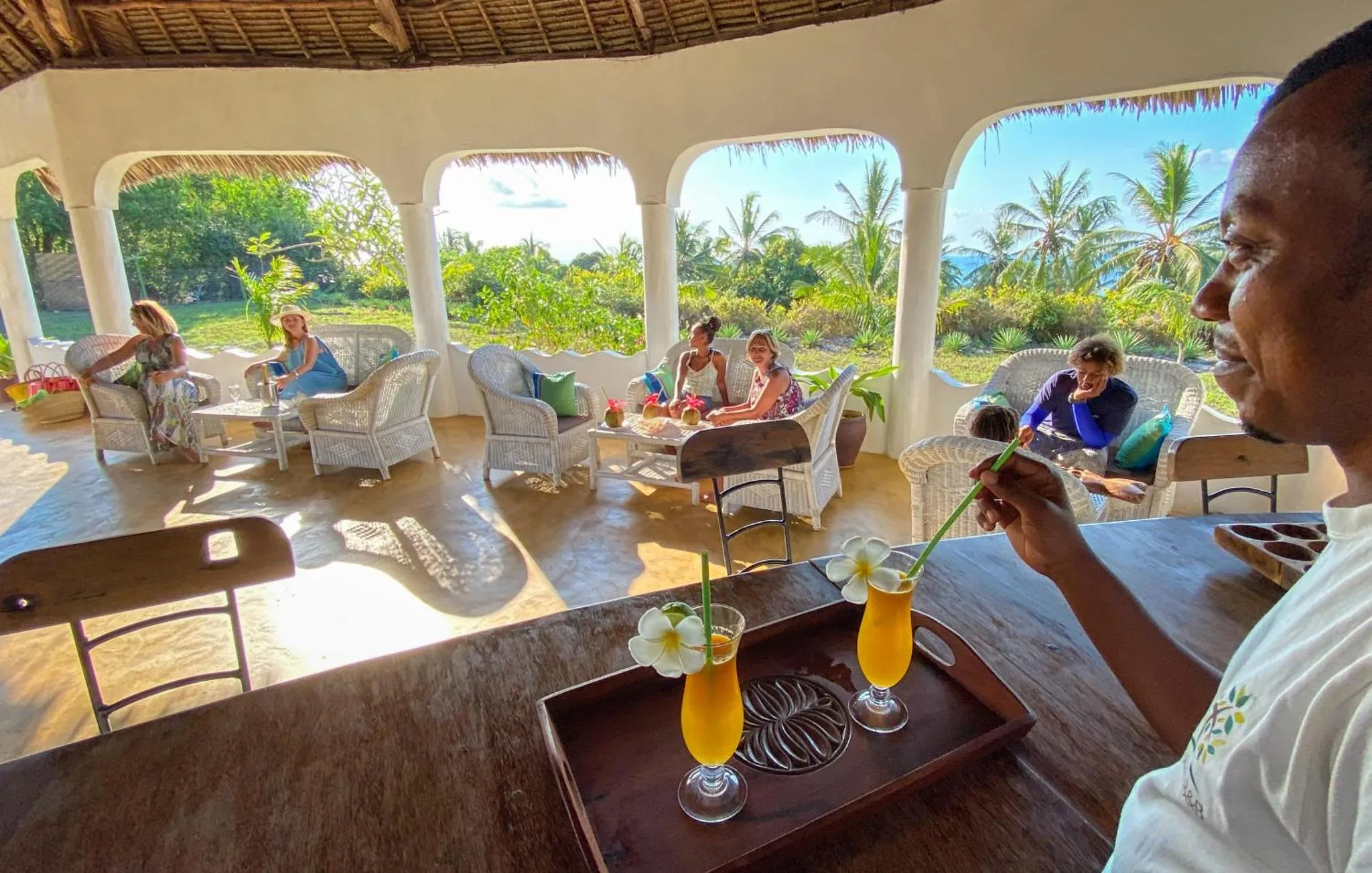 Un homme prépare des boissons sur une terrasse ensoleillée avec des clients assis à des tables.
