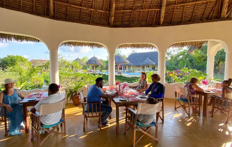 Des convives sont attablés dans une salle à manger ouverte sur une végétation luxuriante et une piscine.