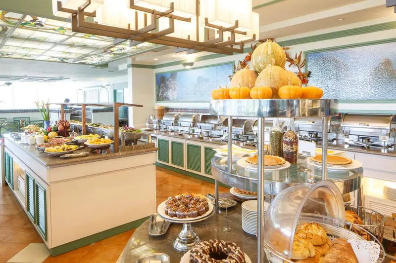Un buffet garni de fruits, pâtisseries et autres mets est présenté dans une salle lumineuse.