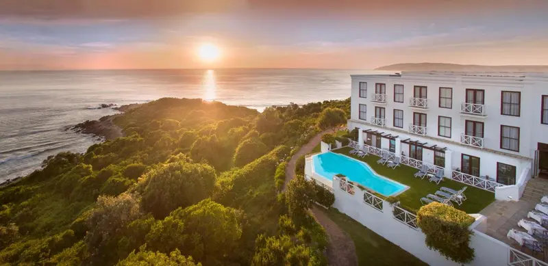 Bâtiment blanc avec piscine et végétation luxuriante au bord de la mer au coucher du soleil.