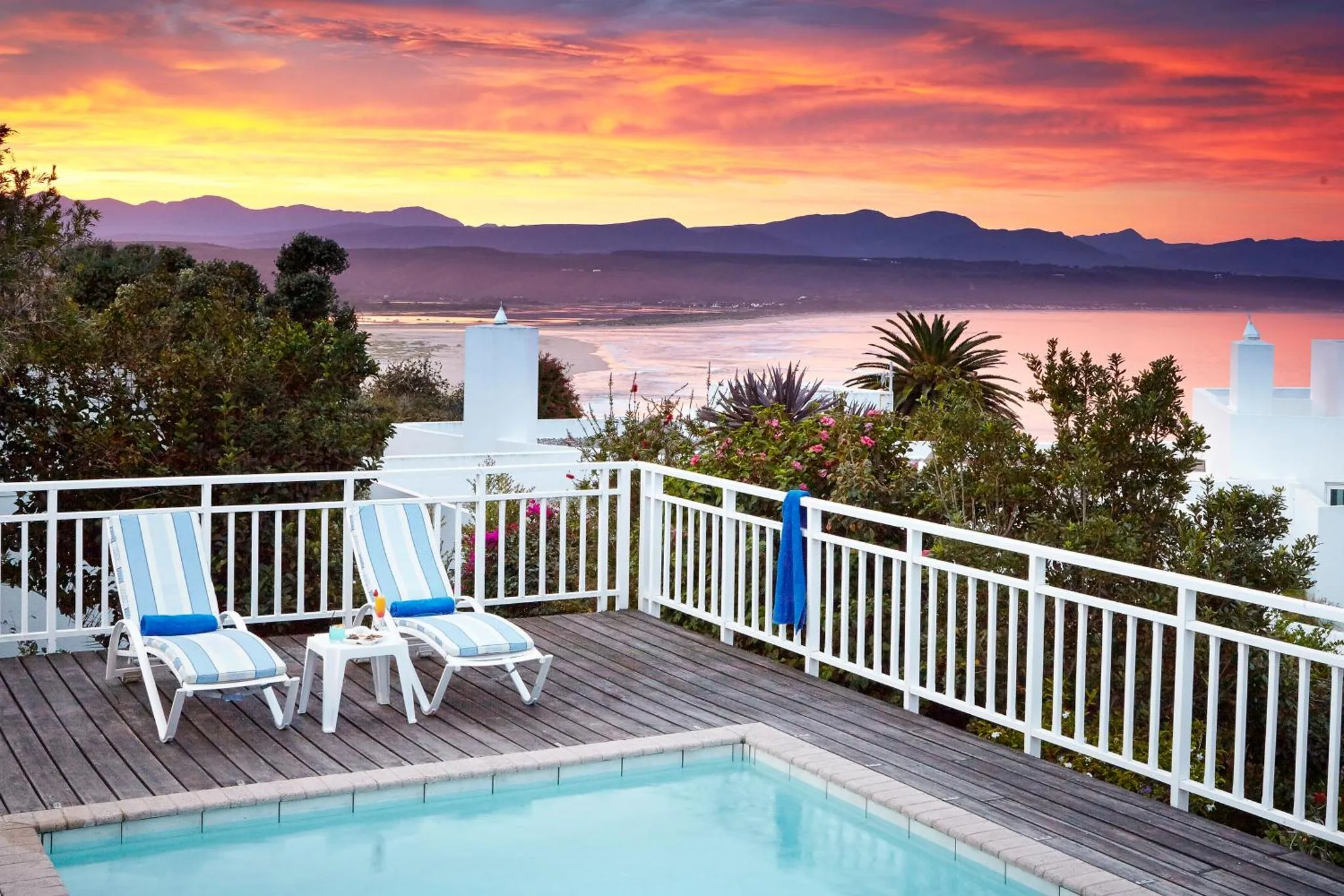 Deux transats bleus invitent à la détente sur une terrasse en bois avec vue sur la mer et un coucher de soleil flamboyant.