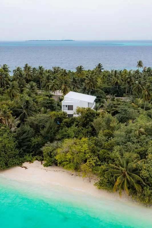 Vue aérienne d'une plage de sable blanc bordée de végétation luxuriante et d'une maison blanche.