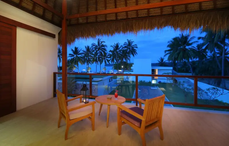 Terrasse aménagée avec mobilier en bois donnant sur une plage et des palmiers au coucher du soleil.
