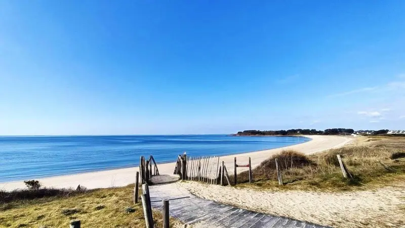 Une plage de sable fin mène vers une mer bleue sous un ciel dégagé.