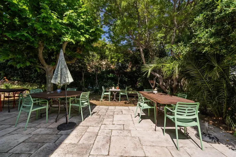 Tables et chaises vertes disposées sur une terrasse pavée ombragée par des arbres.