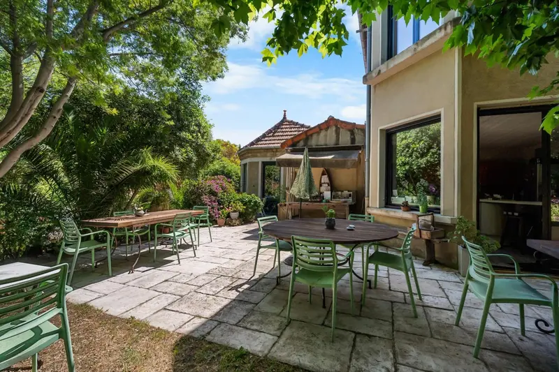 Tables et chaises vertes disposées sur une terrasse pavée dans un jardin luxuriant.