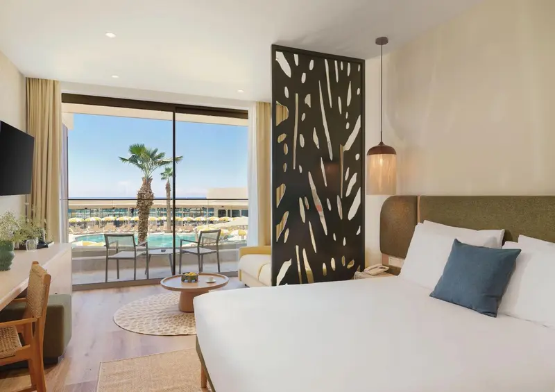 Chambre lumineuse avec un grand baie vitrée donnant sur une piscine et une plage.