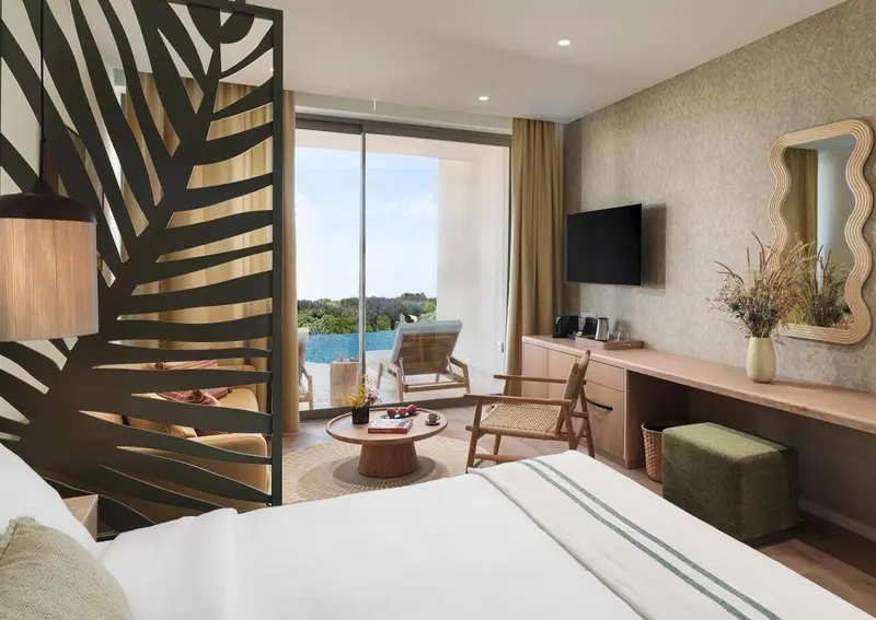 Chambre lumineuse avec un lit blanc et une vue sur une terrasse et la mer.