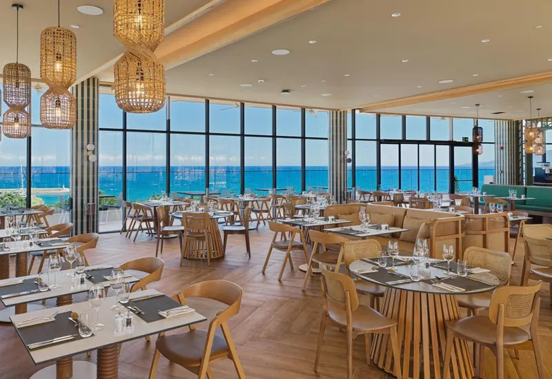 Une salle lumineuse avec de grandes baies vitrées donnant sur l'océan et des tables dressées pour le repas.