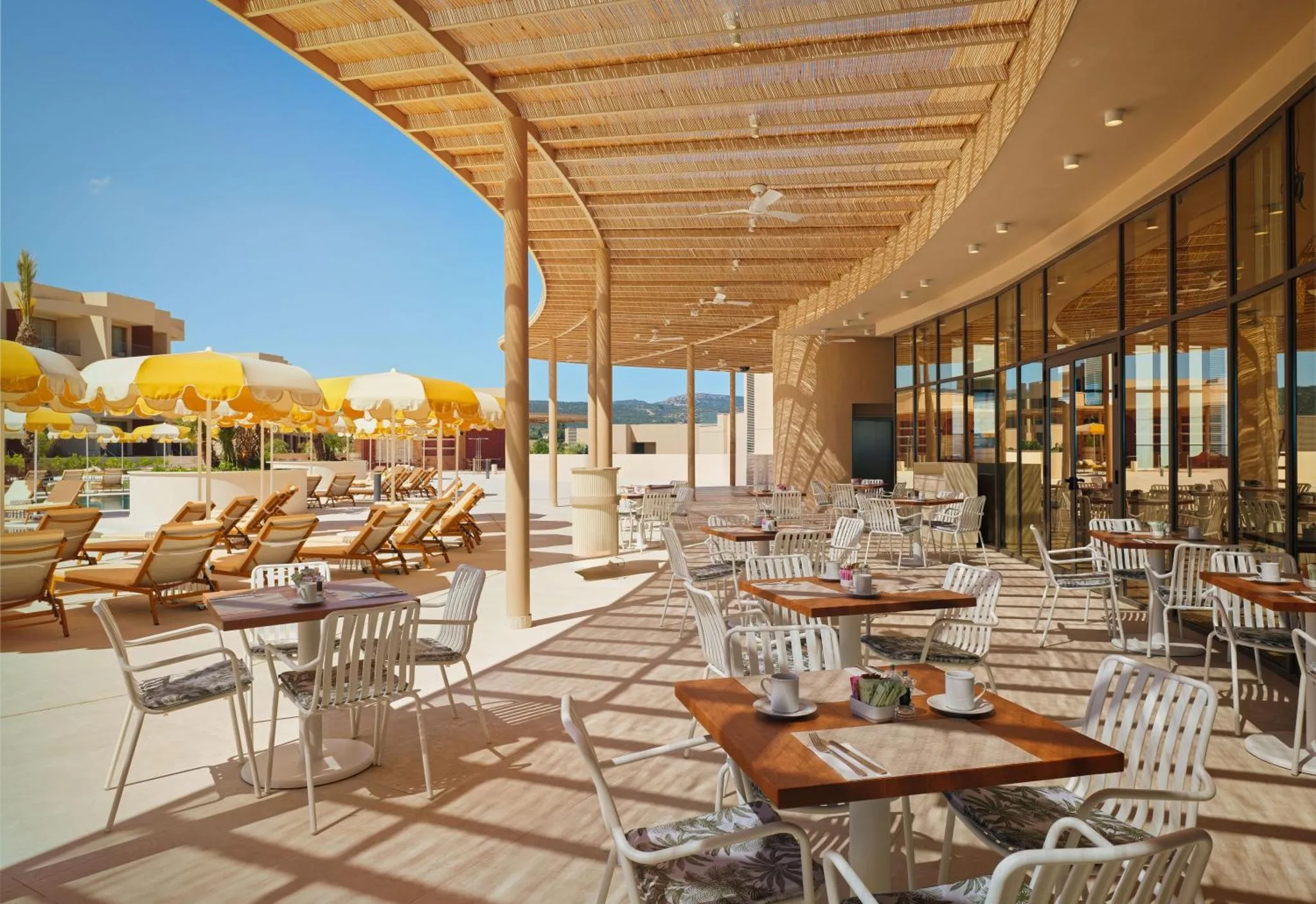 Terrasse ombragée avec tables et chaises, donnant sur une piscine et des transats.