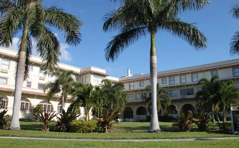 Bâtiment blanc à plusieurs étages entouré de végétation tropicale sous un ciel bleu.