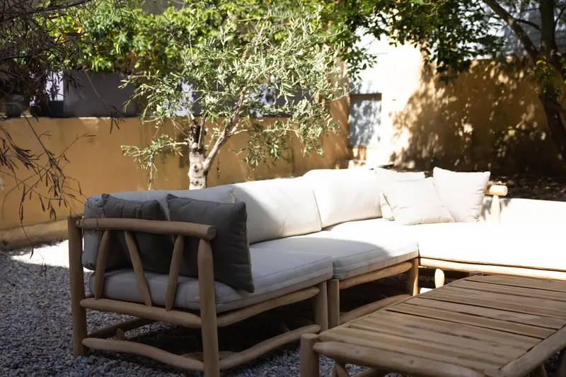 Canapé d'extérieur en bois et coussins blancs dans un jardin ensoleillé.