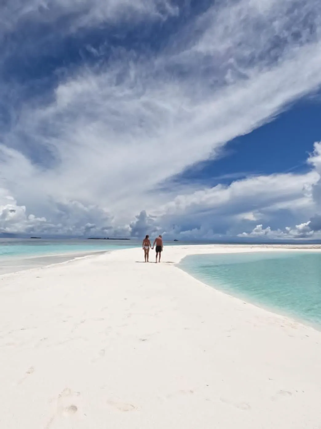 Deux personnes marchent sur une plage de sable blanc bordée d'eau turquoise sous un ciel nuageux.