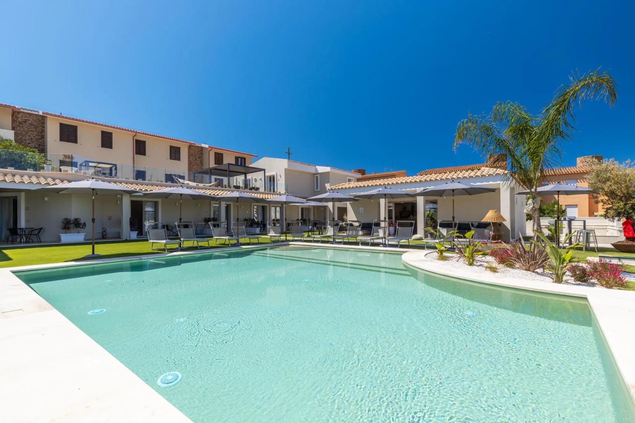 Piscine rectangulaire turquoise bordée d'une terrasse avec transats et parasols, devant un bâtiment aux toits en tuiles.