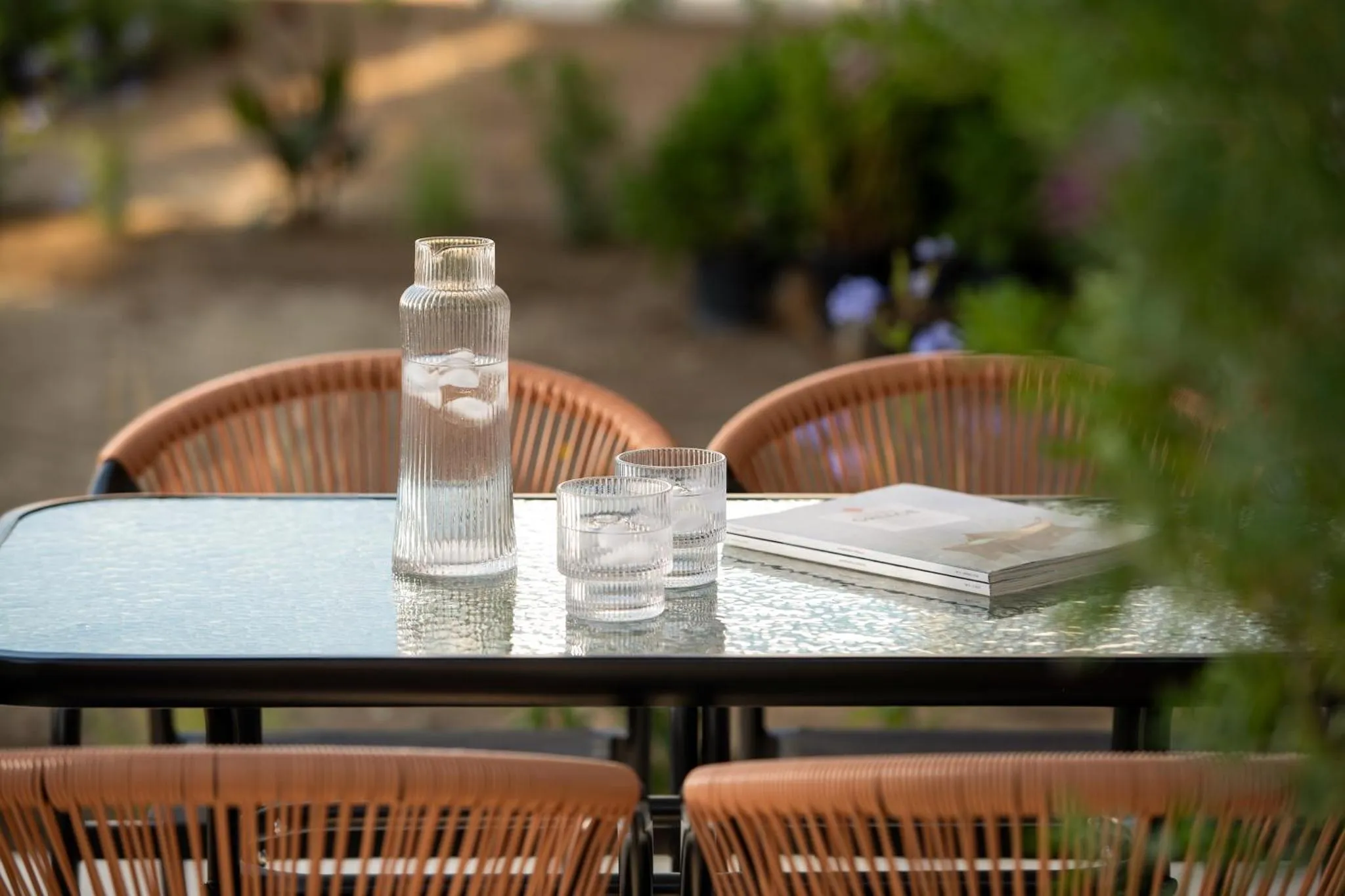 Une table basse en verre entourée de chaises en rotin accueille une carafe d'eau et des magazines.