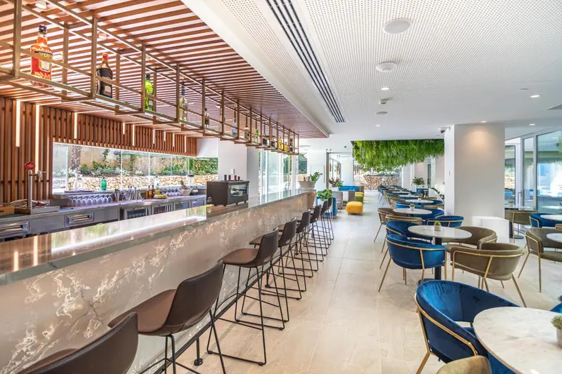 Un long comptoir de bar moderne avec des tabourets, devant un mur végétal et une salle à manger avec des tables et des chaises.