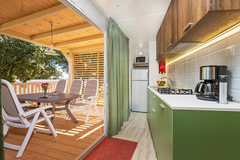 Une cuisine verte s'ouvre sur une terrasse en bois avec une table et des chaises.