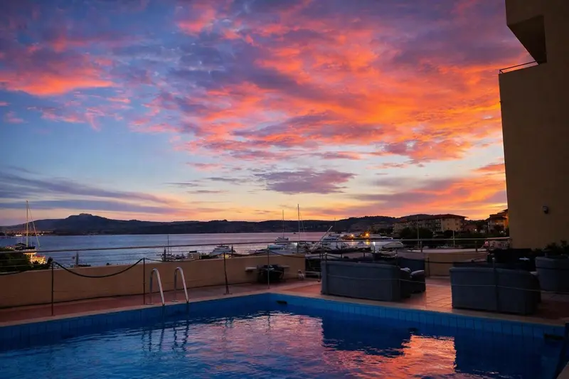 Une piscine au premier plan offre une vue sur un port illuminé et un ciel flamboyant au coucher du soleil.
