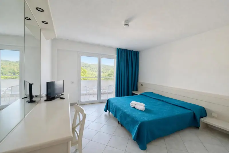 Chambre lumineuse avec un lit turquoise et une vue sur la végétation.