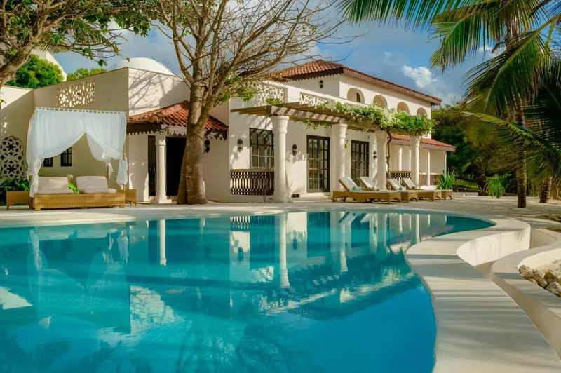 Vue d'une piscine turquoise devant un bâtiment blanc à colonnes et une terrasse ombragée.