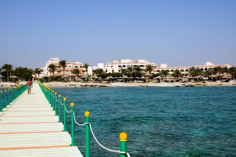 Un ponton en bois mène vers un complexe hôtelier blanc au bord de la mer turquoise.