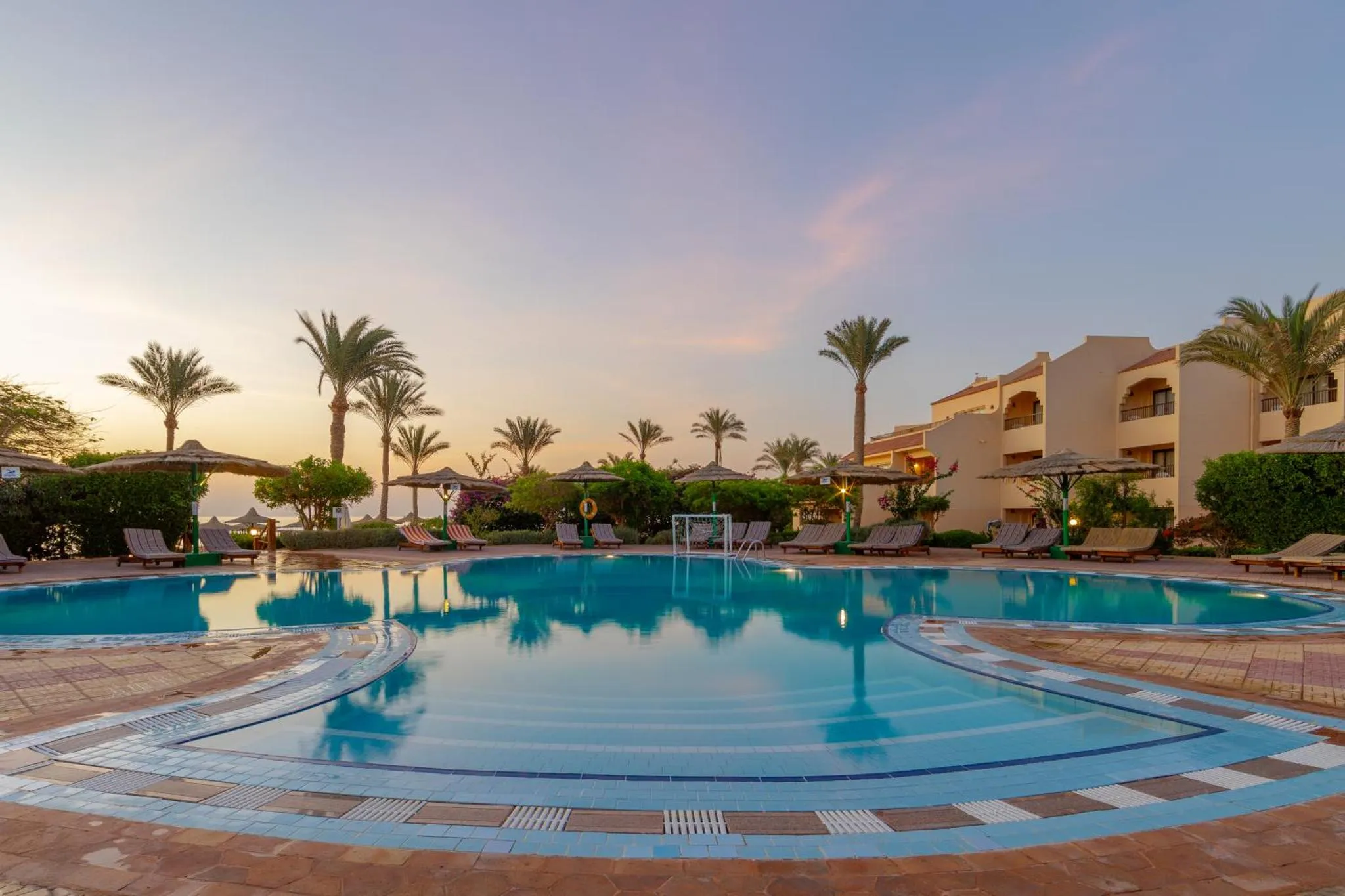 Piscine turquoise bordée d'une terrasse avec transats et palmiers au coucher du soleil.