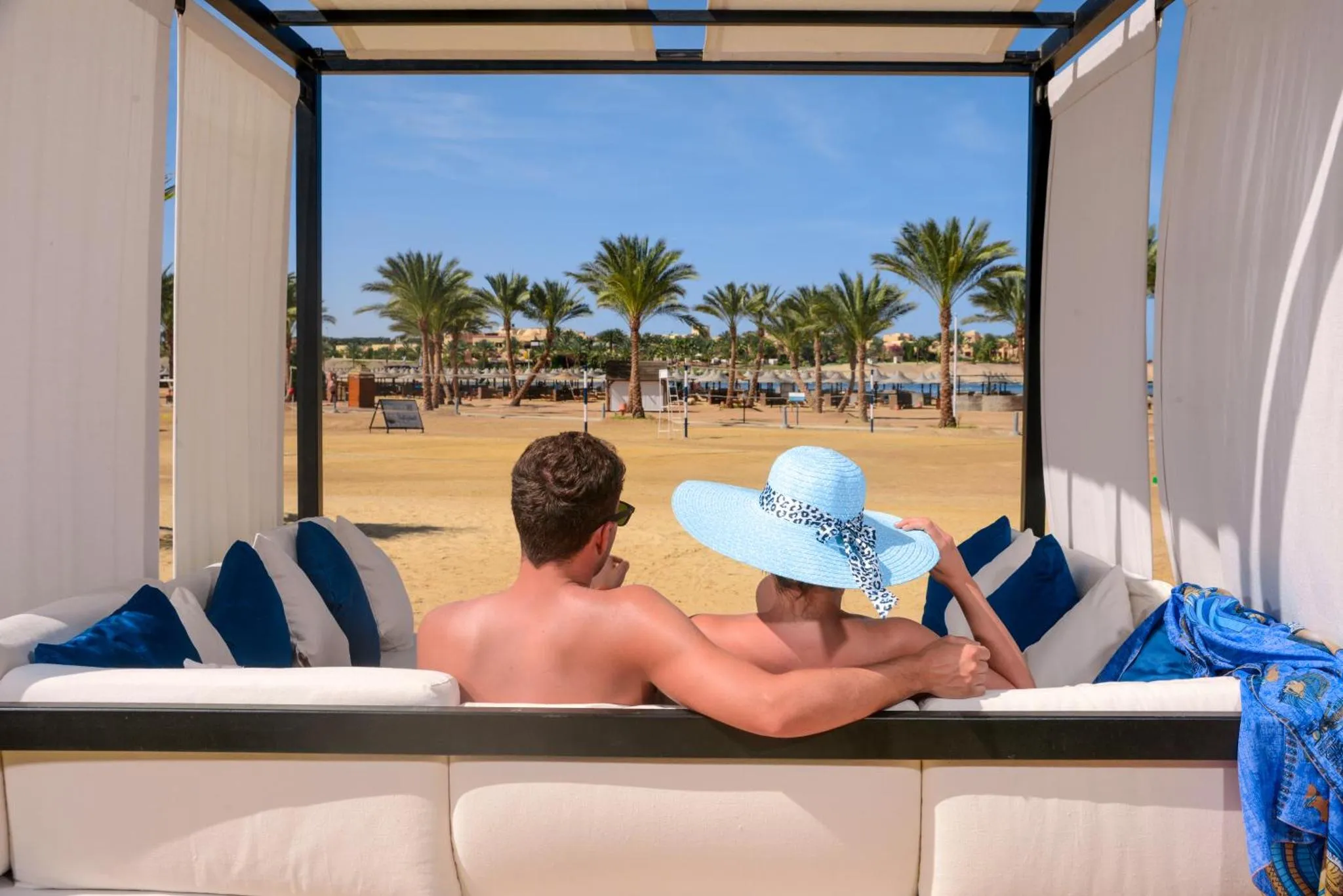 Deux personnes se relaxent sur des coussins blancs dans un espace ombragé face à une plage de sable.