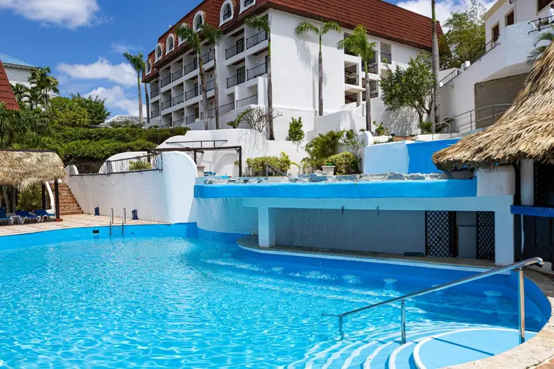 Une piscine turquoise bordée d'une terrasse ensoleillée devant un bâtiment blanc.
