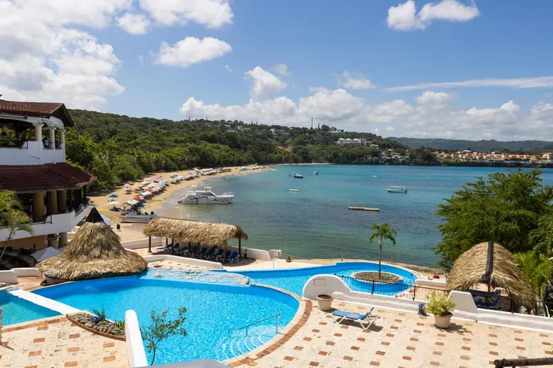 Vue aérienne d'une piscine avec transats surplombant une plage de sable fin et une baie turquoise.