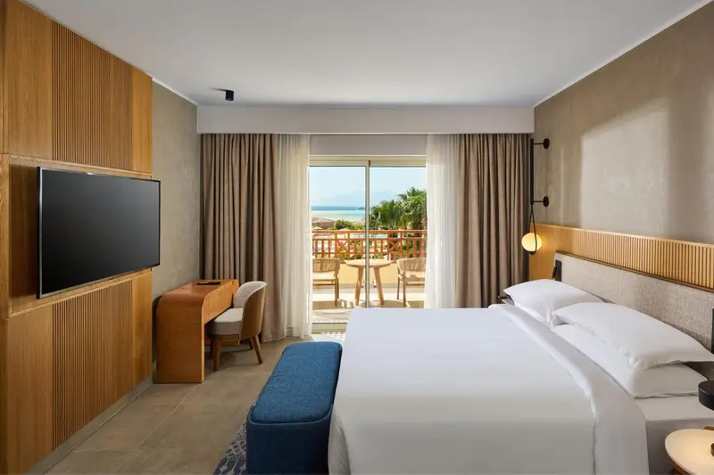 Chambre lumineuse avec un grand lit et une vue sur la mer depuis un balcon.