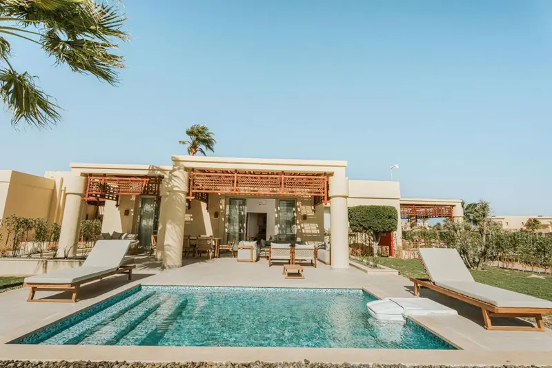 Petite piscine devant une terrasse ombragée et un bâtiment beige.