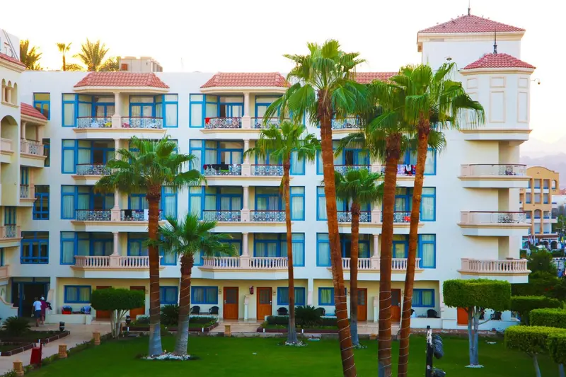 Bâtiment blanc à plusieurs étages avec des balcons et des palmiers devant un jardin verdoyant.
