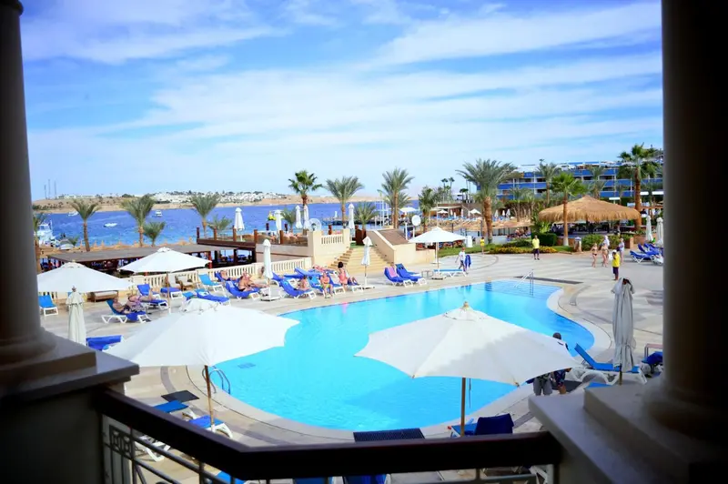 Vue aérienne sur une piscine entourée de transats et de parasols, avec la mer en arrière-plan.