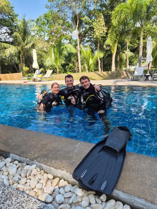 Trois personnes souriantes en combinaison de plongée se tiennent dans une piscine turquoise.