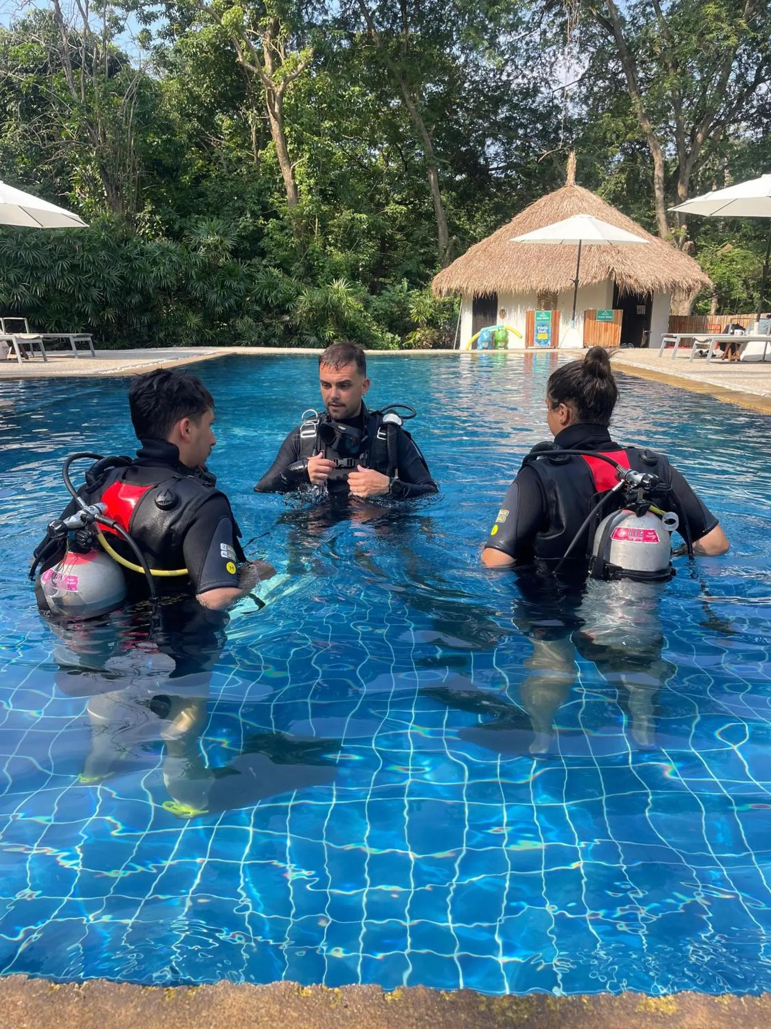 Trois personnes en équipement de plongée discutent au bord d'une piscine.