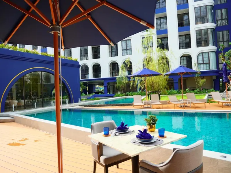 Une table est dressée au bord d'une piscine turquoise, avec un parasol bleu au premier plan.