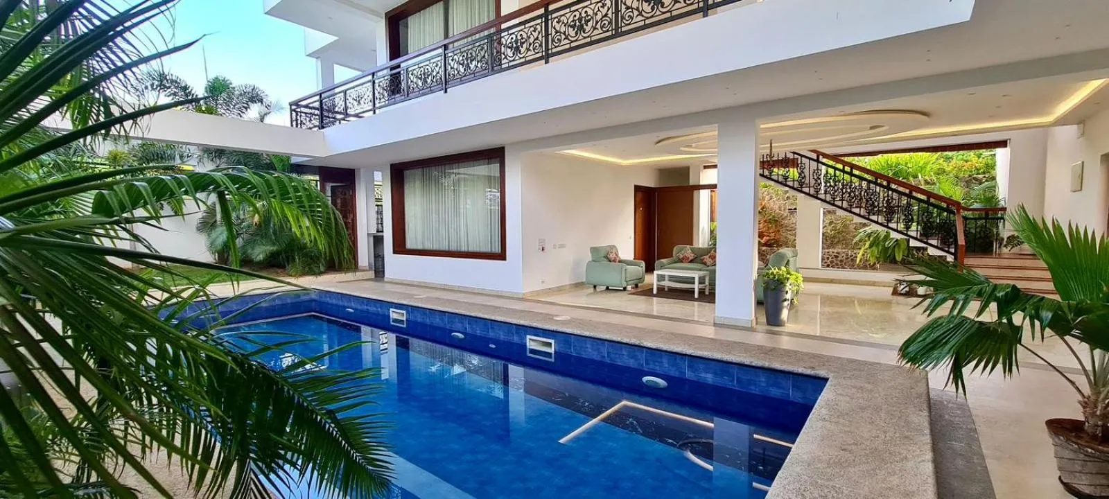 Une piscine turquoise est bordée d'une terrasse et d'une végétation luxuriante devant un bâtiment blanc.