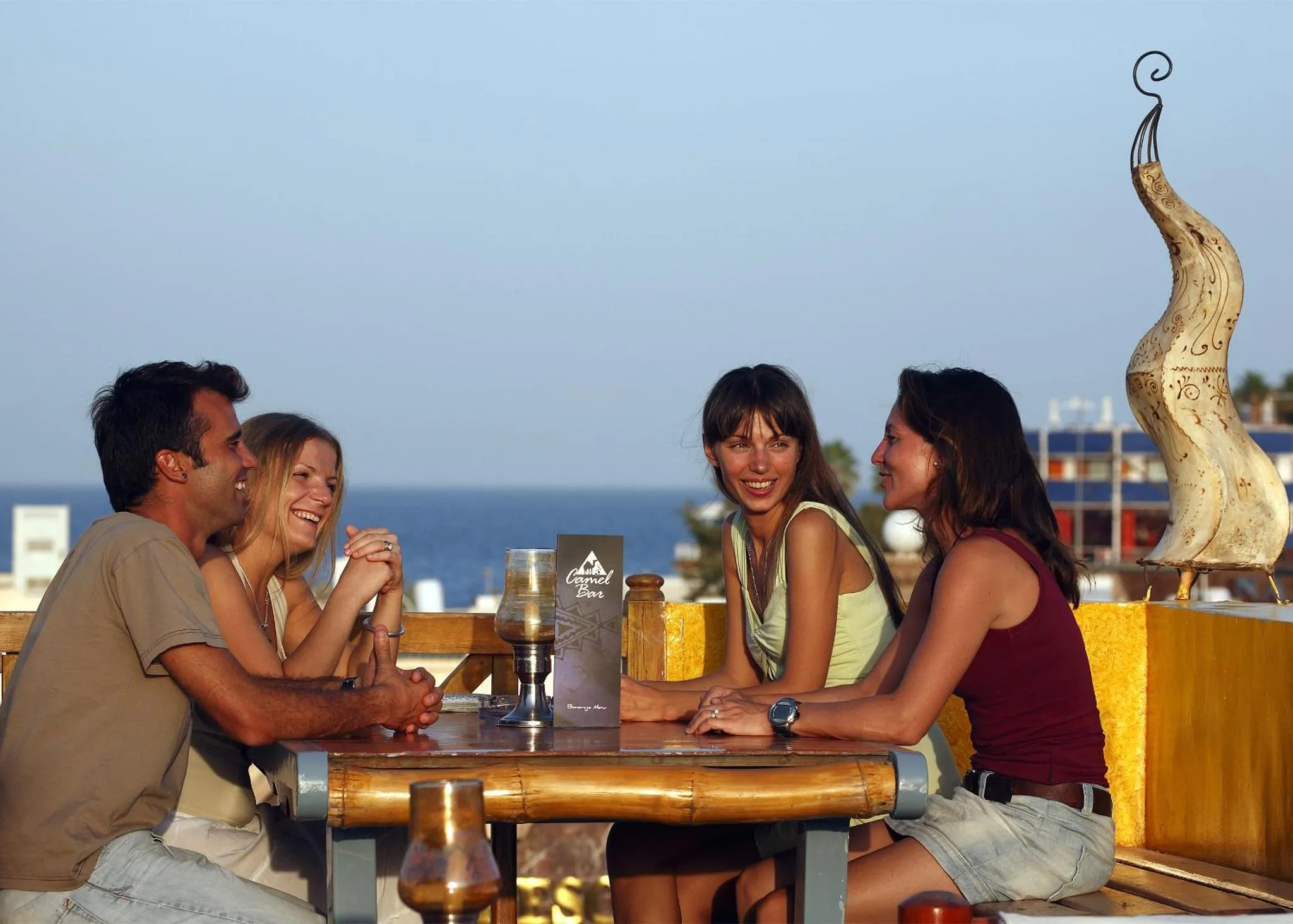 Quatre personnes sourient en sirotant des boissons à une table en bois sur une terrasse ensoleillée.