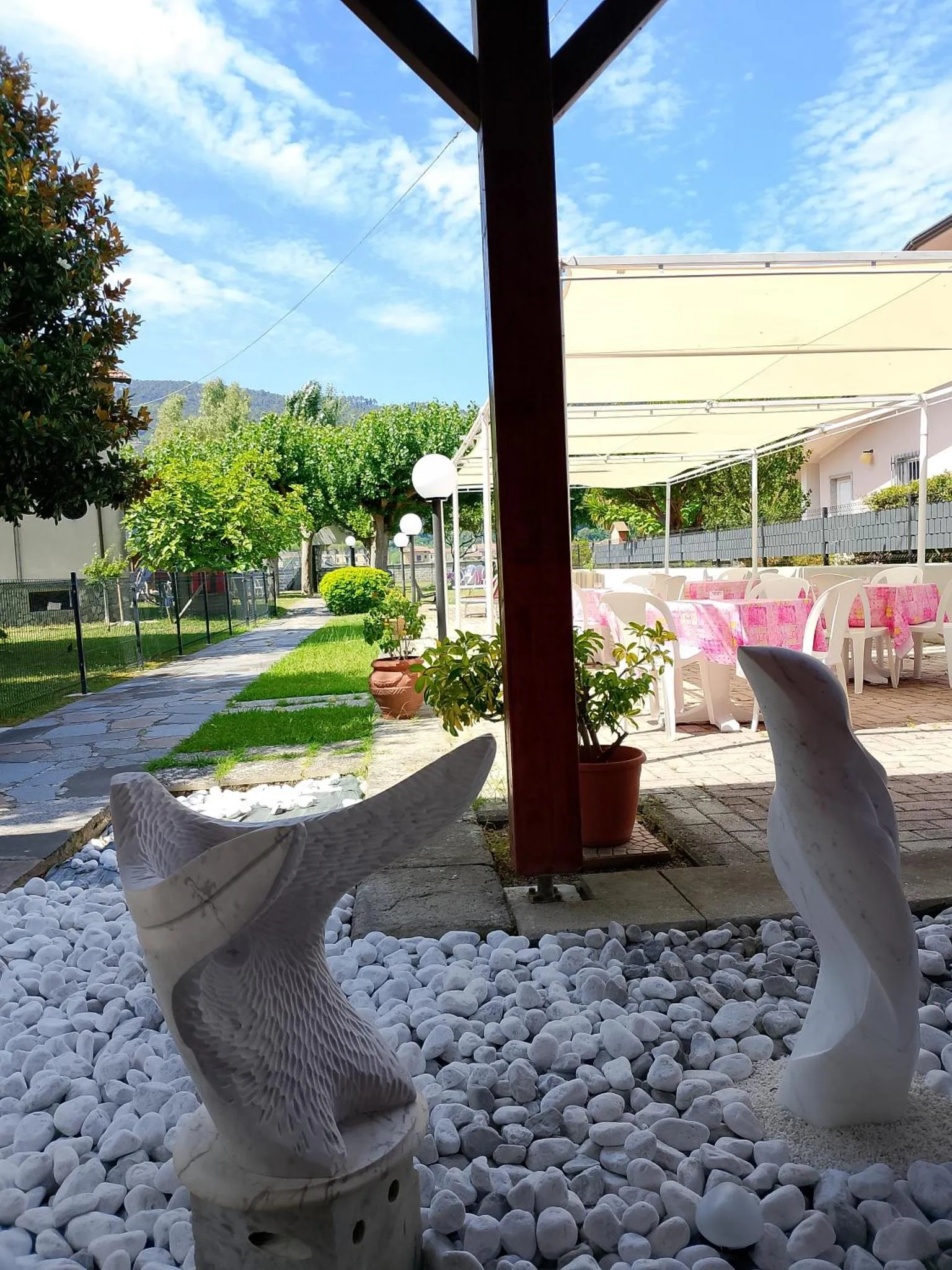 Terrasse ombragée avec tables et chaises, décorée de statues de dauphins et de plantes.