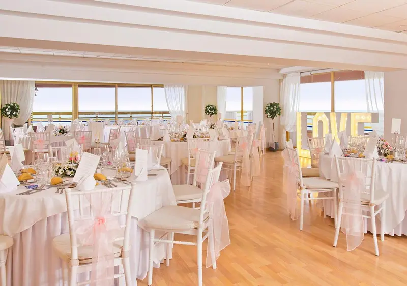 Une salle lumineuse est mise en scène pour un banquet avec des tables rondes nappées et décorées de fleurs.