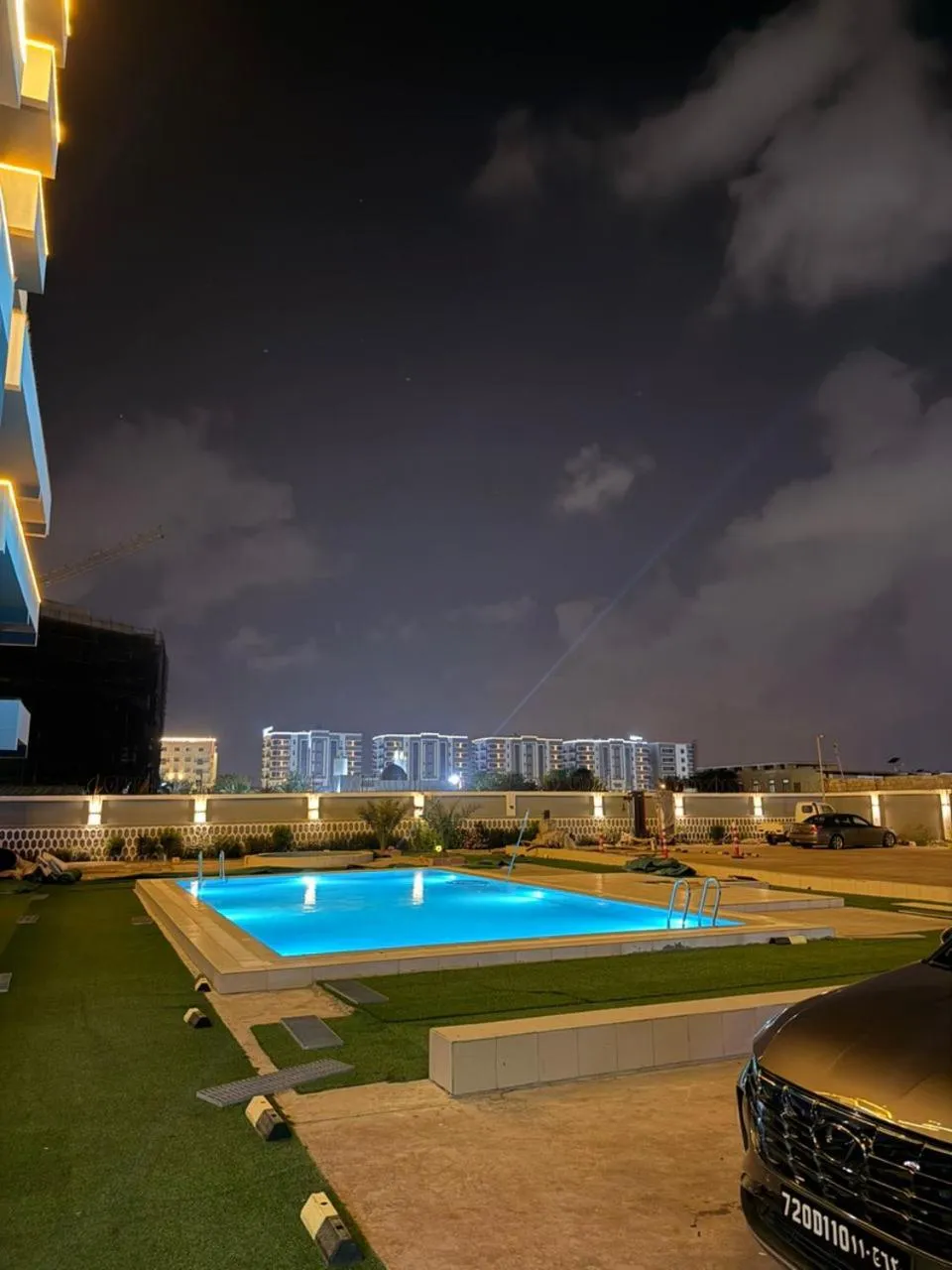 Une piscine illuminée au crépuscule avec une pelouse verdoyante et des bâtiments en arrière-plan.