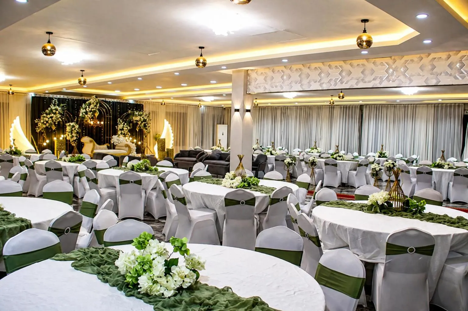 Une grande salle lumineuse est aménagée pour un événement avec des tables rondes nappées et décorées de fleurs.