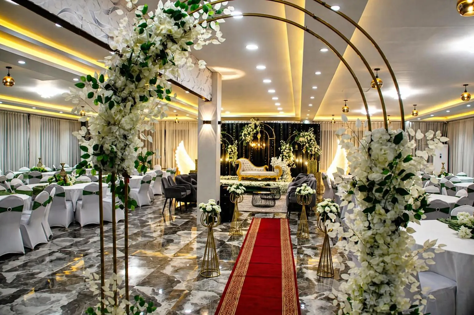 Une salle de réception décorée de fleurs et d'arches dorées accueille des tables dressées pour un événement.